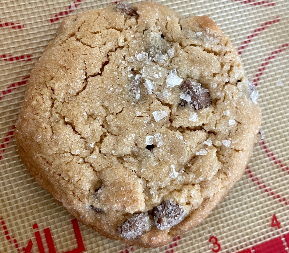 A large, baked chocolate chip and peanut butter cookie. There is flaky sea salt on top. The viewer is looking directly over the cookie. It sits on a red and white silicone mat.