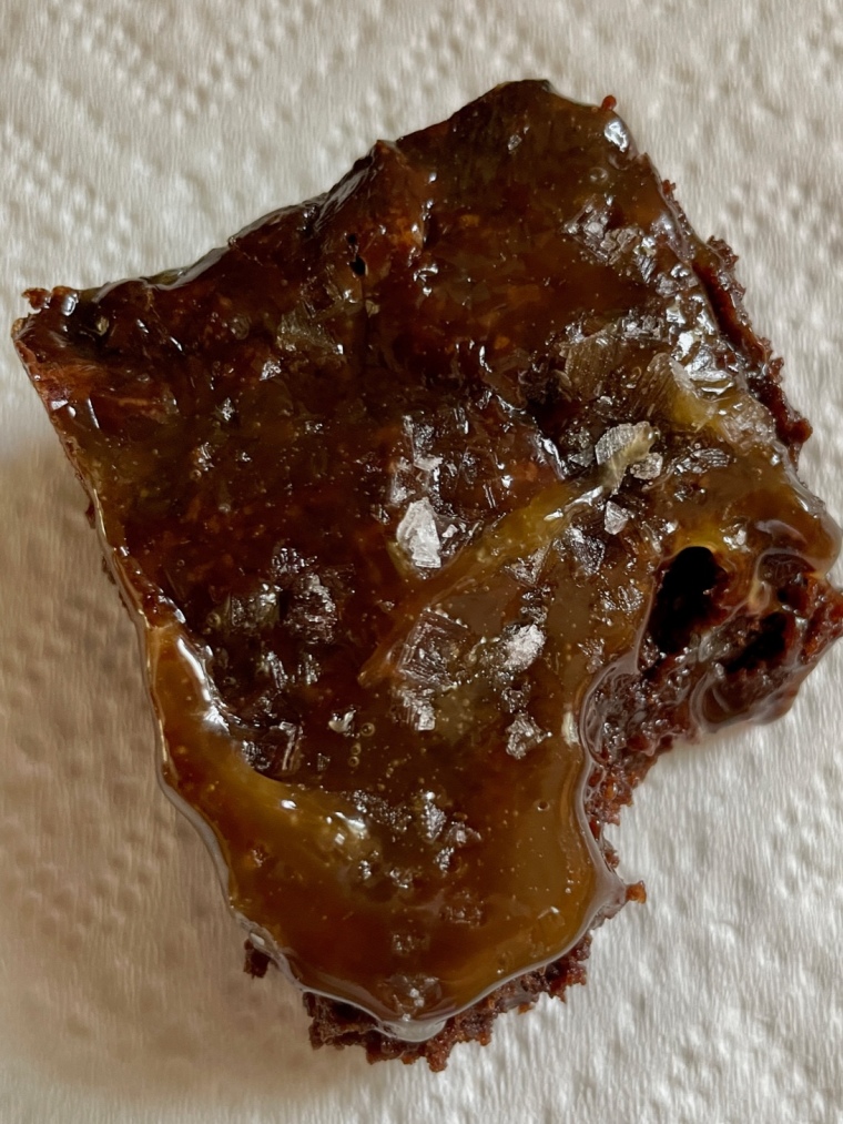 One brownies cut from the sheet, with a bite taken out of it. The shot is a close up to see the texture of the brownie, caramel, and salt.